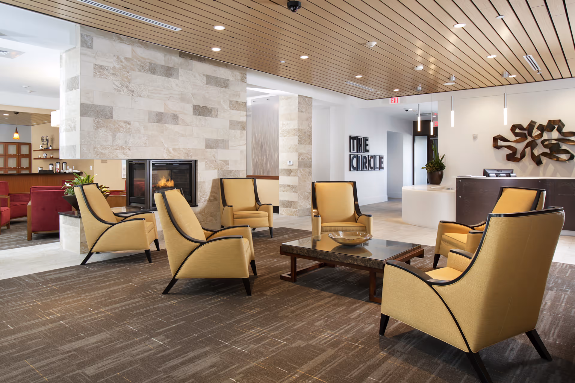 A modern senior living facility common area with six beige armchairs arranged around a low rectangular coffee table. There is a stone fireplace in the background with a fire burning inside. The ceiling is wooden with recessed lighting, and there is a reception desk to the right with a decorative wall sculpture above it. A sign on the wall reads 'THE CIRCLE'.
