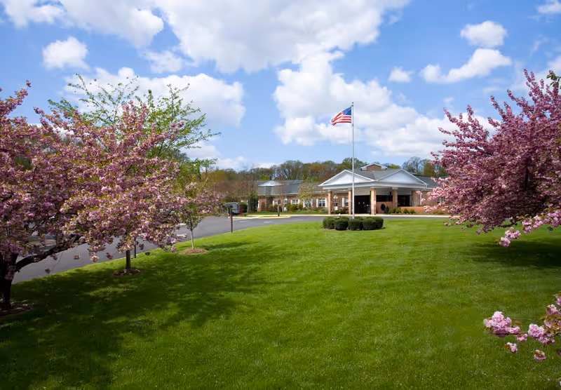 A well-maintained lawn with blooming pink cherry blossom trees on either side, leading to the entrance of a brick building with a white portico. An American flag flies on a flagpole in the center of the lawn under a partly cloudy blue sky.