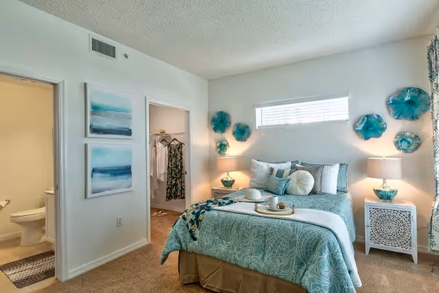 A neatly arranged bedroom with a bed covered in teal and white bedding, decorative pillows, and a tray with a cup and hat. The room has beige carpet, two nightstands with matching lamps, teal wall decorations above the bed, a small horizontal window, and an open doorway leading to a walk-in closet and a bathroom with a toilet visible.