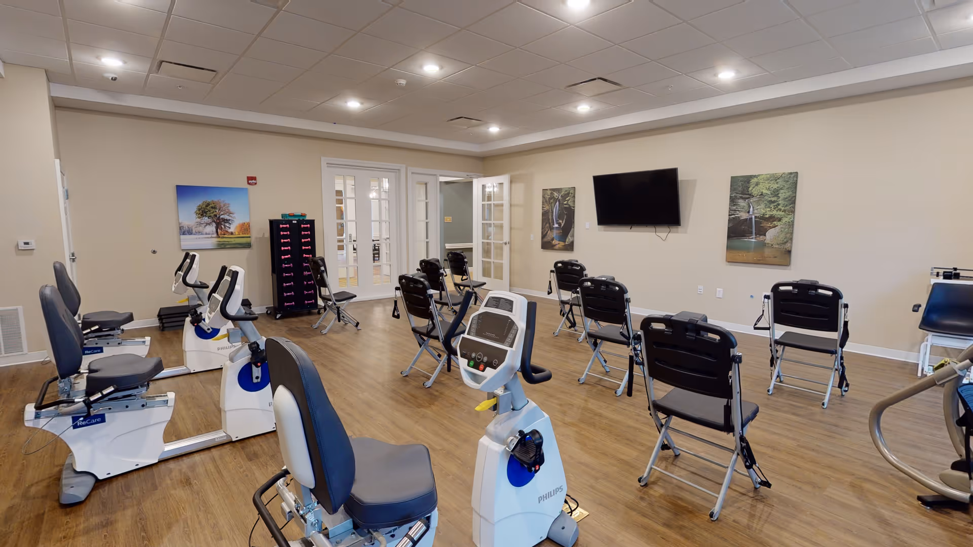 A spacious exercise room with several stationary recumbent bikes and chairs arranged facing a wall-mounted flat screen TV. The room has wood flooring, beige walls, recessed ceiling lights, and framed nature artwork on the walls. French doors lead to another room in the background.