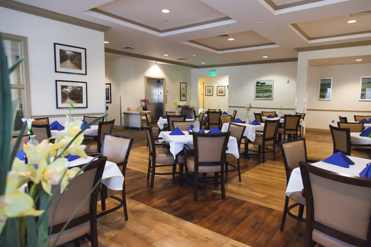 A dining room in a senior living facility with multiple tables covered with white tablecloths and blue folded napkins. Each table has a small vase with flowers. The room has wooden flooring, beige walls with framed artwork, and recessed ceiling lights. There is an elevator and seating area visible in the background.