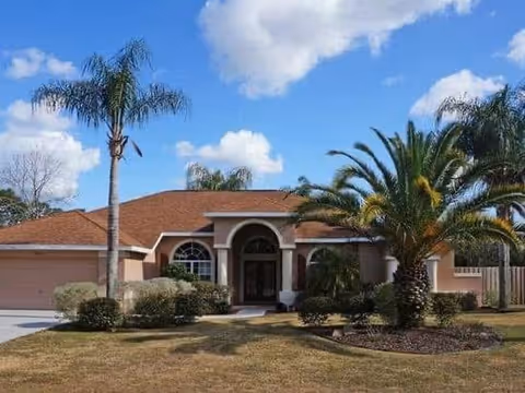 Single-story residential building with a brown roof, arched entrance, and large windows, surrounded by palm trees and landscaping under a partly cloudy blue sky.