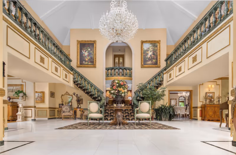 Spacious and elegant interior of a senior living facility with a grand double staircase featuring green railings. A large crystal chandelier hangs from the high ceiling. Two framed floral paintings are mounted on the wall above the staircase. Below, a decorative table with a large floral arrangement is flanked by two green upholstered chairs on a patterned rug. The room is decorated with plants, statues, and classic furniture, with a polished white tile floor.