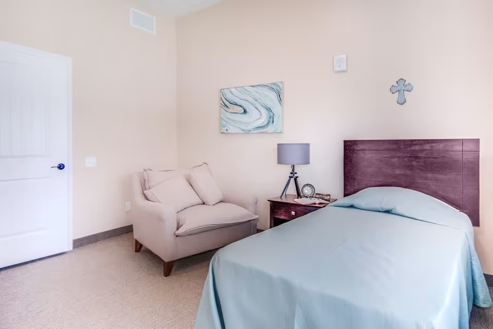 A simple bedroom with a single bed covered in a light blue bedspread, a wooden headboard, a small wooden nightstand with a lamp, clock, and glasses on it, a beige armchair with cushions, a white door, and light-colored walls decorated with a blue and white abstract painting and a small cross.