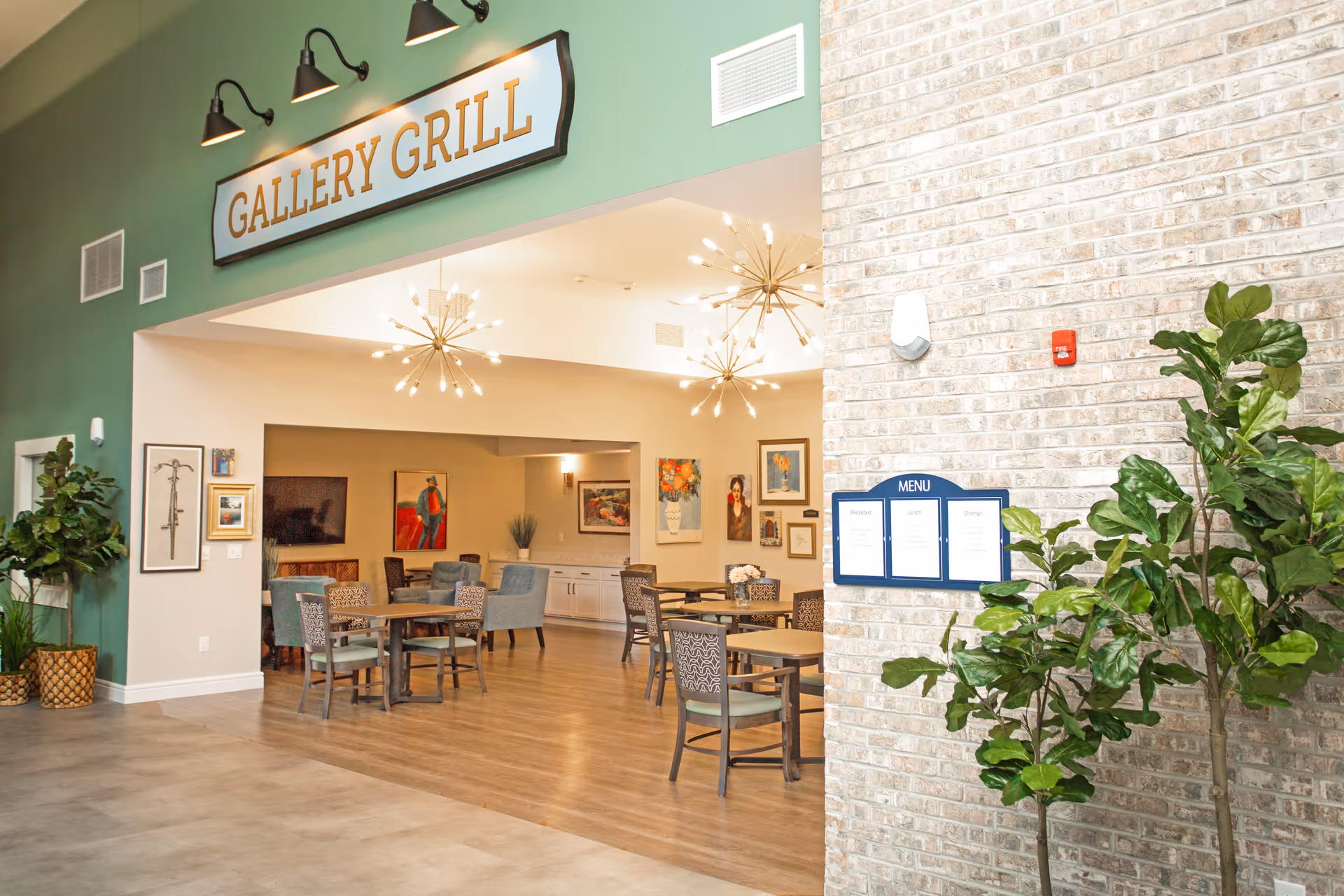 Interior view of a dining area named Gallery Grill with several tables and chairs arranged on a wooden floor. The walls are decorated with framed artwork, and modern light fixtures hang from the ceiling. There are green plants near a brick wall that has a menu displayed on it.