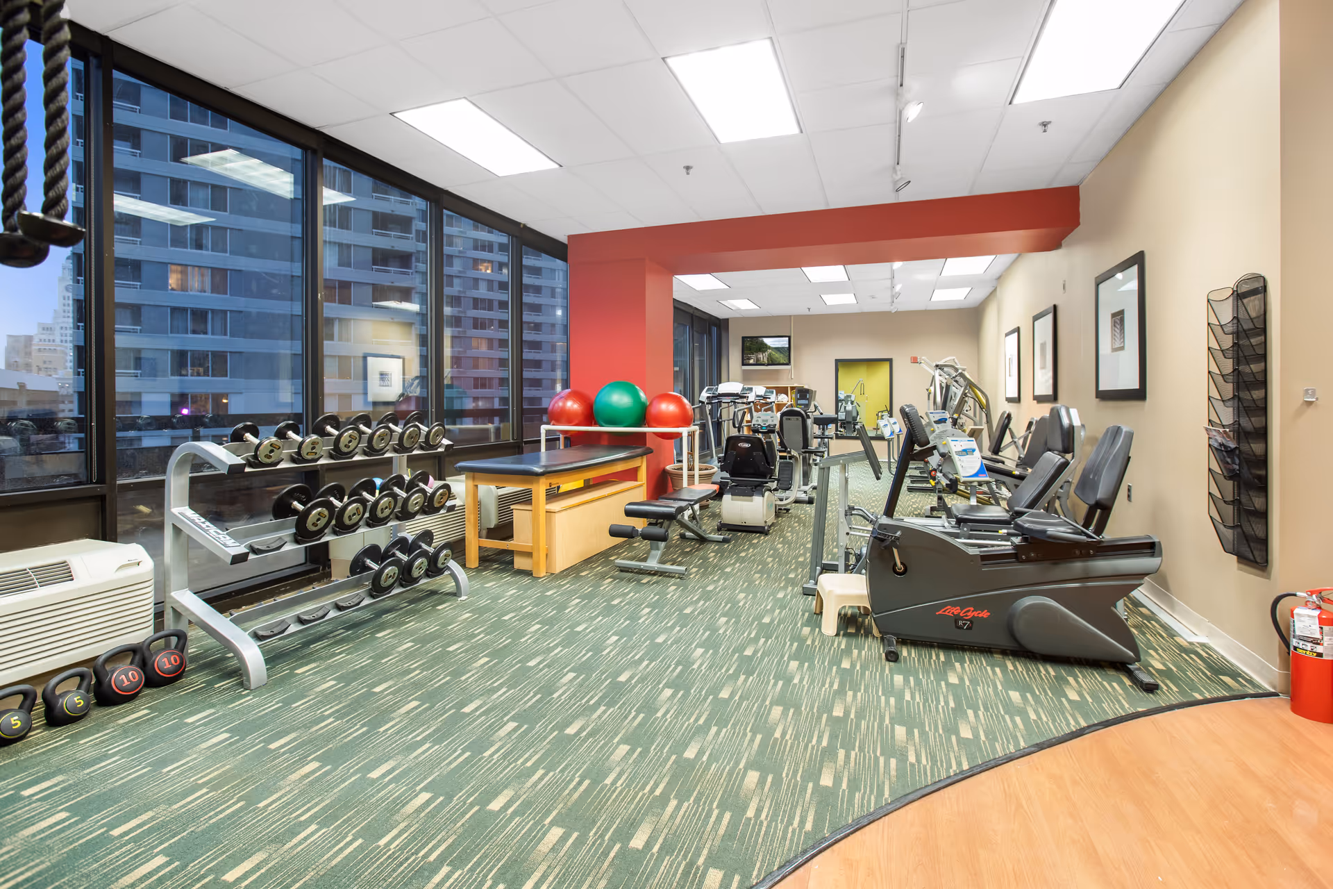 A fitness room in a senior living facility with various exercise equipment including dumbbells, kettlebells, exercise balls, stationary bikes, and weight machines. Large windows show a cityscape outside, and the room has green patterned carpet and beige walls with framed pictures.