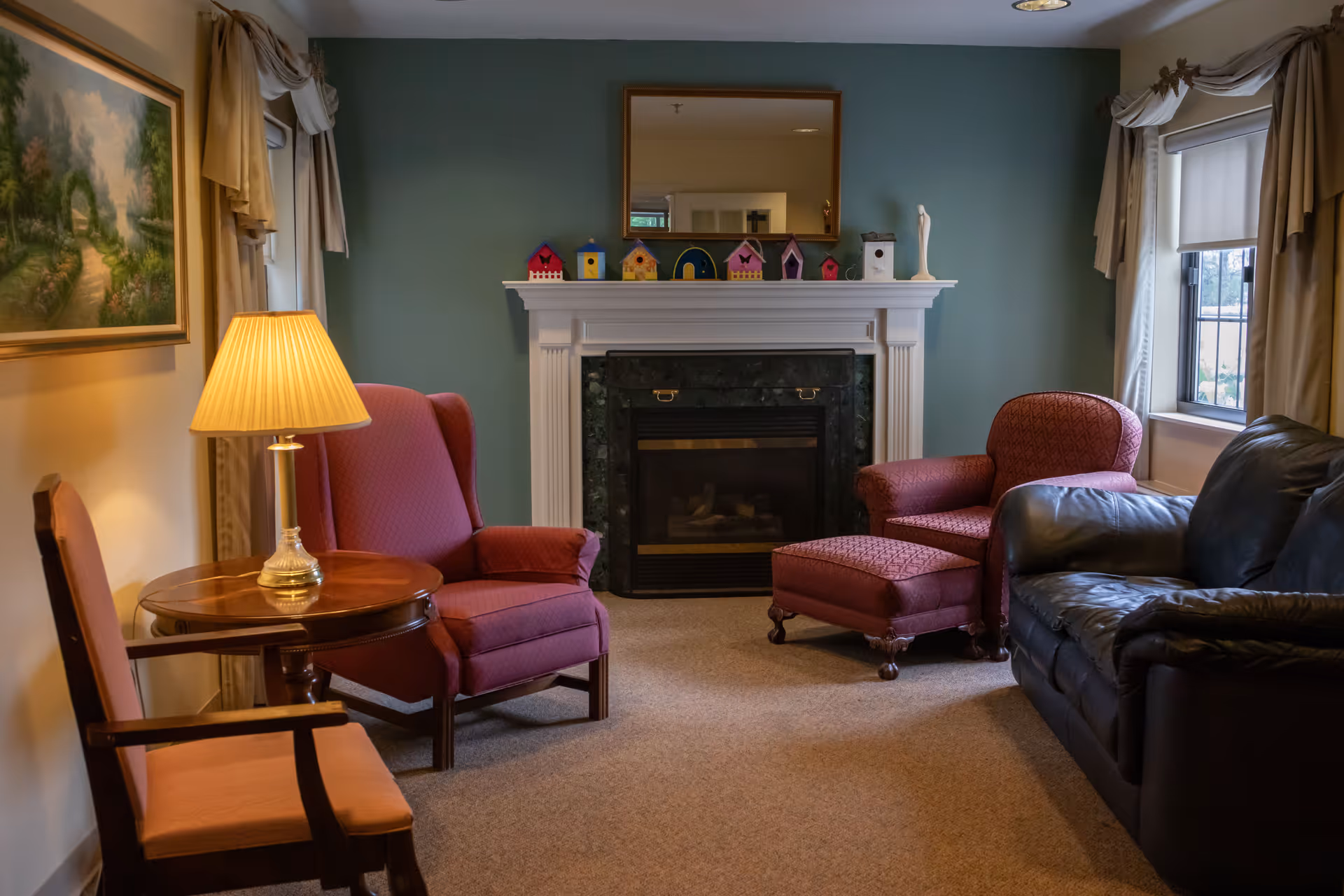 A cozy living room with a green accent wall featuring a white fireplace decorated with colorful birdhouses and a small statue. The room has a large mirror above the fireplace, a black leather sofa, two red upholstered armchairs, one with a matching ottoman, a wooden side table with a lamp, and a window with beige curtains and a white roller shade.