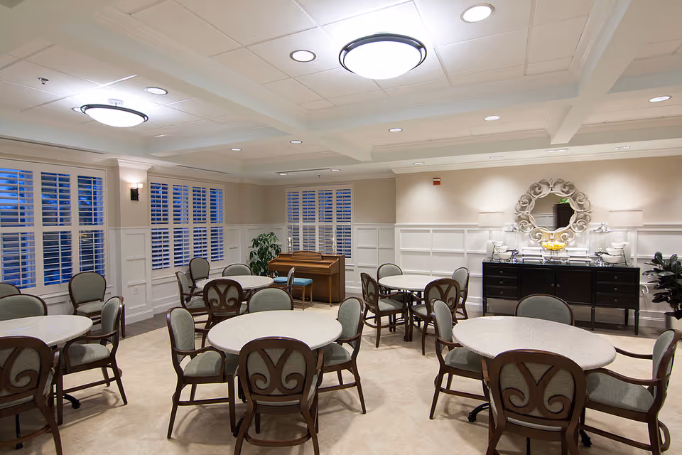 A well-lit dining room with round tables and cushioned chairs arranged neatly. The room features large windows with white shutters, a piano in the corner, and a black sideboard with decorative items and a round ornate mirror above it. The ceiling has recessed lighting and two large circular light fixtures.