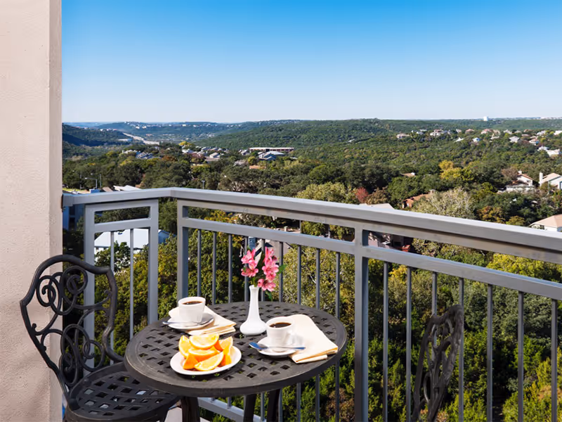 A balcony with a round black metal table and two matching chairs overlooking a green hilly landscape. On the table are two cups of coffee, a plate with orange slices, a small stack of books, a napkin with a spoon, and a white vase with pink flowers.