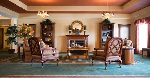 A cozy living room area with two upholstered armchairs facing a wooden coffee table and an electric fireplace. The room features a green carpet, two wooden bookshelves with decorative items, a round mirror above the fireplace, and two chandeliers hanging from the ceiling. There are large windows with curtains on the right side and some plants adding greenery to the space.