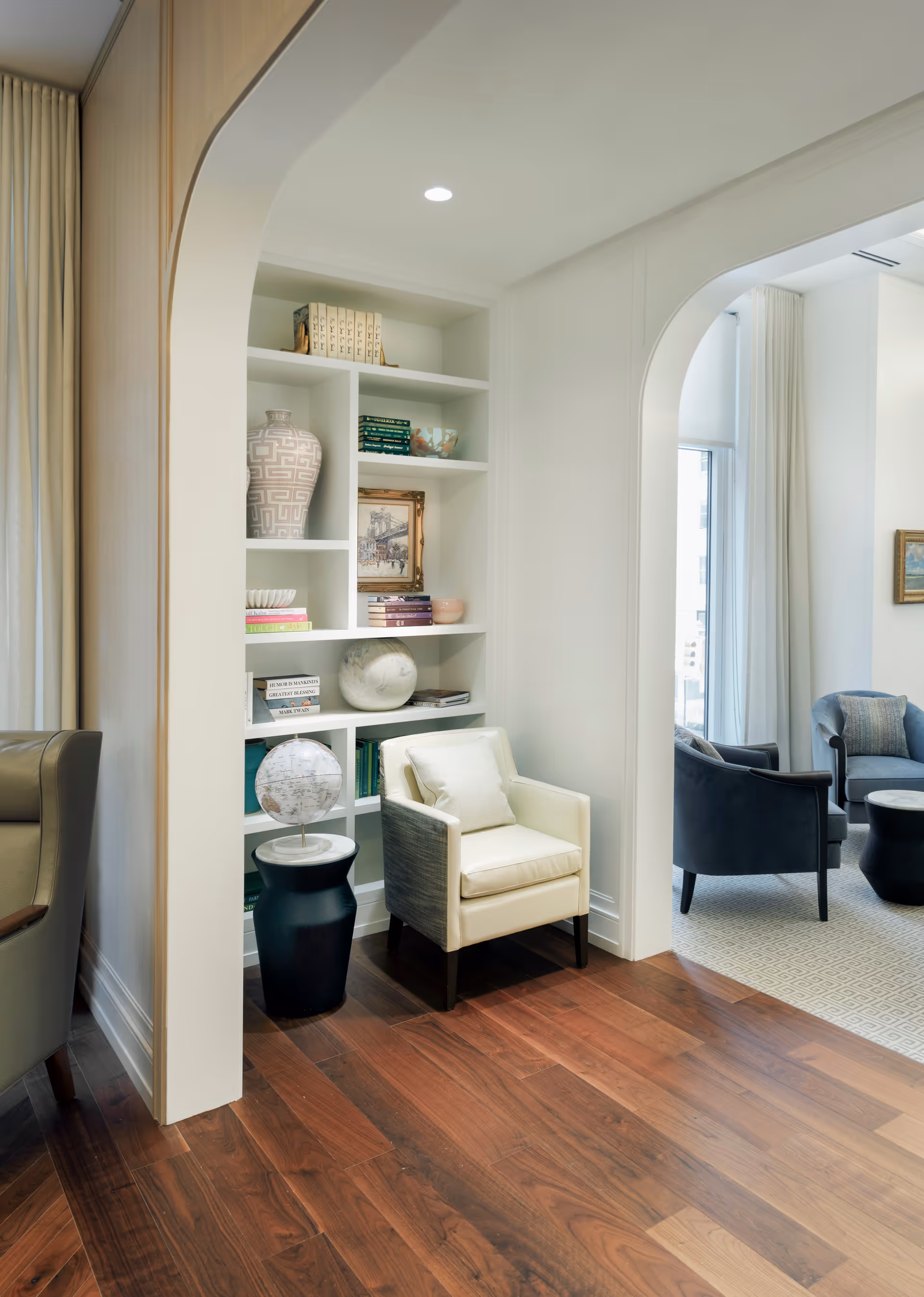A cozy interior space featuring a white armchair with a cushion placed next to a black side table holding a globe. Behind the chair is a built-in white bookshelf filled with books, decorative vases, a framed picture, and other ornaments. The room has wooden flooring and white walls with an arched doorway leading to another seating area with blue armchairs and a small round table near large windows with white curtains.