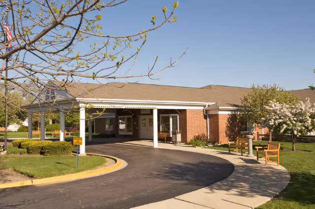 Exterior view of a single-story brick building with a covered entrance driveway. There are green lawns, trees with budding leaves, and wooden benches along a curved sidewalk near the entrance under a clear blue sky.