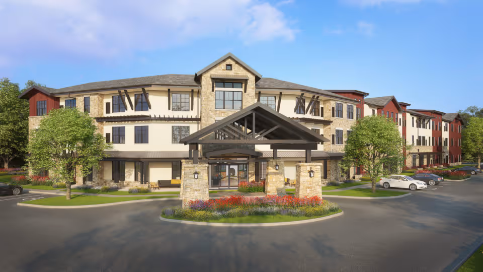 Exterior view of a large, three-story senior living facility building with stone and beige walls, multiple windows, and a covered entrance with stone pillars. The building is surrounded by trees, landscaped flower beds, and a parking area with several cars. The sky is clear and blue.