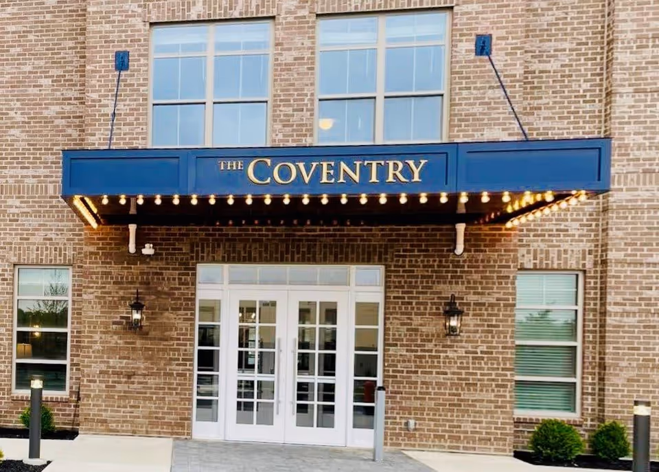 Entrance of The Coventry building with a blue awning displaying the name in gold letters, surrounded by brick walls, two windows, and glass double doors below the awning.