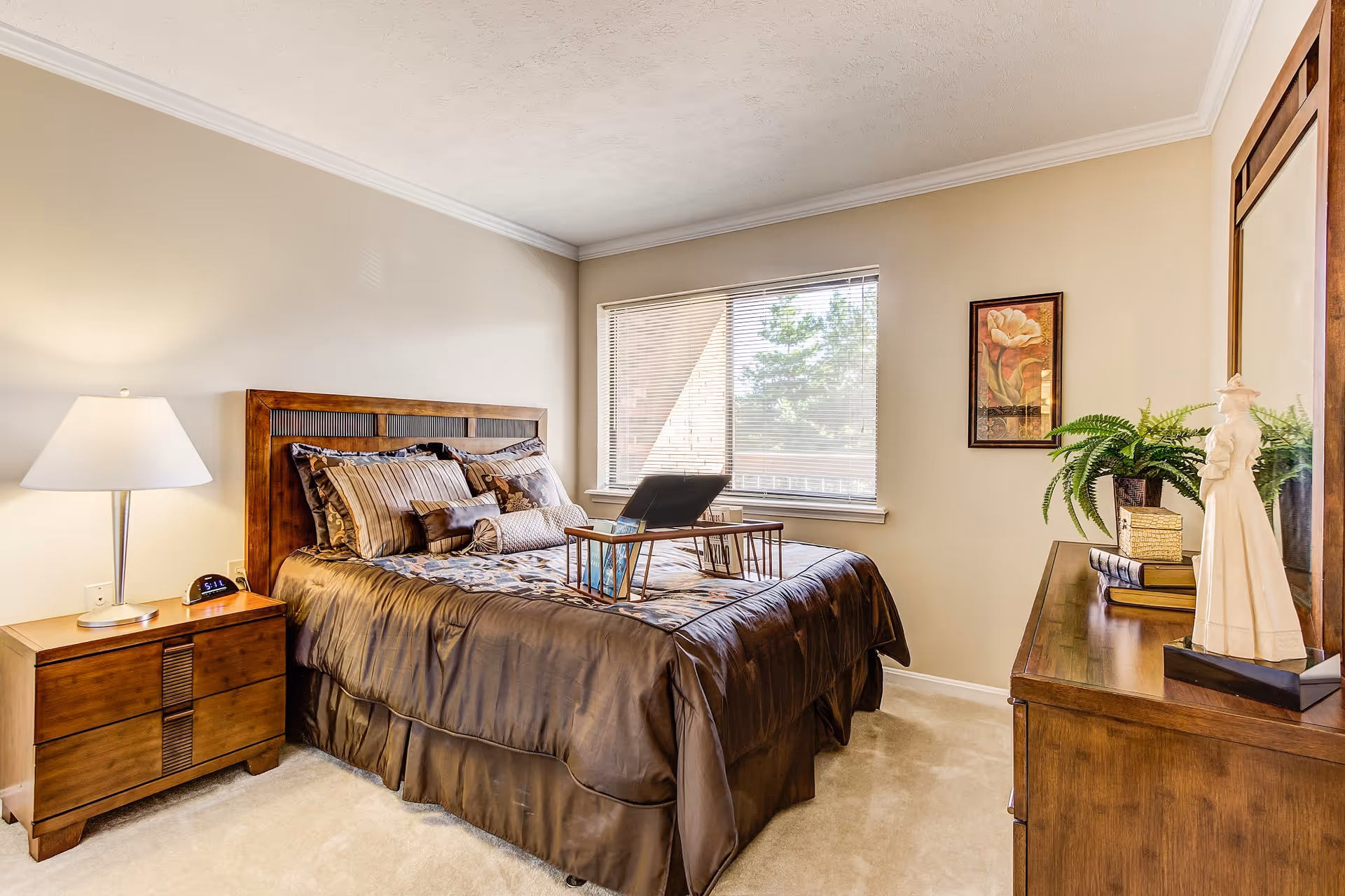 A cozy bedroom with a wooden bed frame and a neatly made bed with brown and beige bedding. A wooden nightstand with a lamp and an alarm clock is beside the bed. There is a window with blinds letting in natural light. A wooden dresser with decorative items, including a plant and a figurine, is on the right side of the room. A framed floral artwork hangs on the wall.