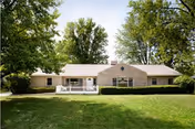 Single-story residential building with beige siding and a white front porch railing, surrounded by green grass and large leafy trees under a clear sky.
