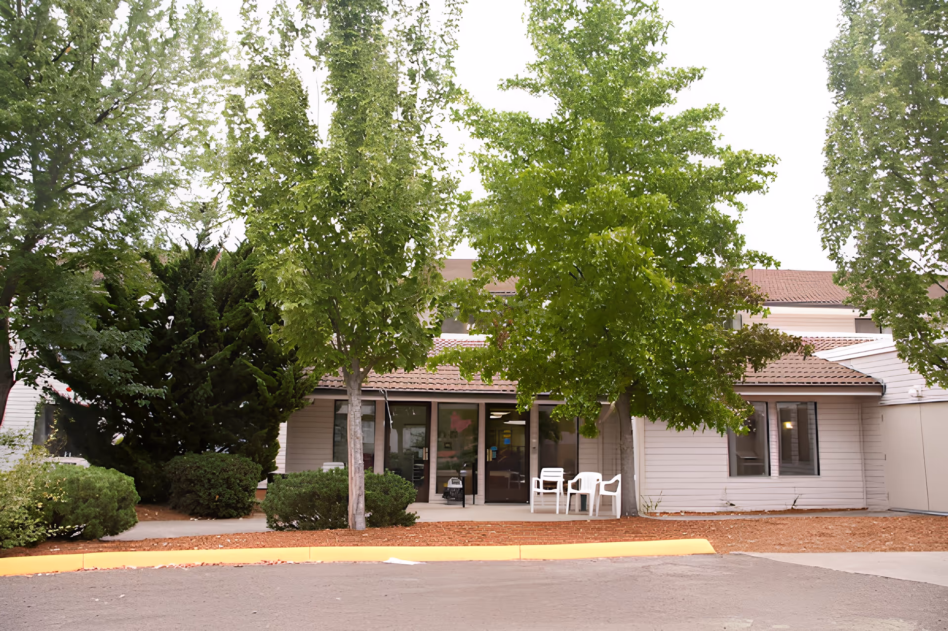 Exterior view of Churchill Estates Retirement Community building entrance with trees and shrubs in front, two white chairs on the porch, and a paved driveway.
