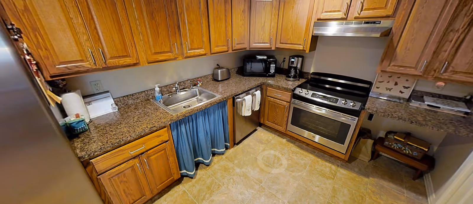 A kitchen with wooden cabinets, granite countertops, a stainless steel sink, a dishwasher with two white towels hanging on the handle, a stainless steel oven and stove, a microwave, a coffee maker, a toaster, and a paper towel holder. The floor is tiled and there is a blue curtain under the sink area.