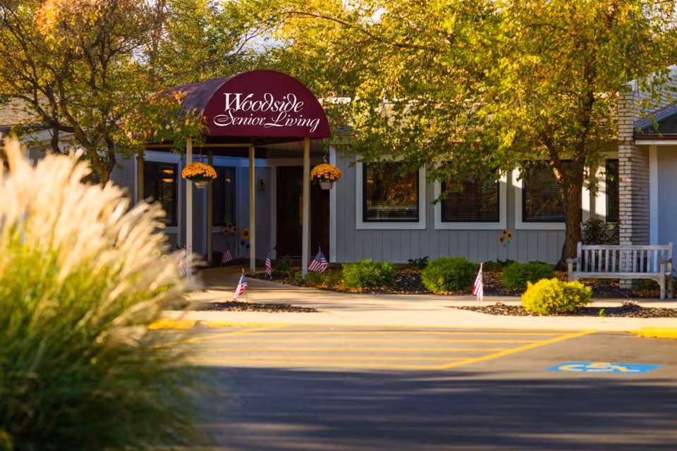 Entrance of Woodside Senior Living facility with a maroon awning displaying the facility name, surrounded by trees, bushes, small American flags, and a bench near the building.