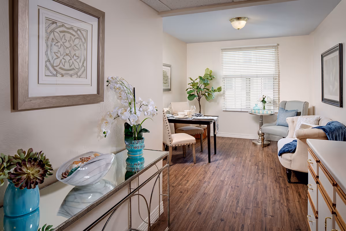A cozy and well-lit living and dining area in a senior living facility. The room features wood flooring, a small dining table set for two with beige upholstered chairs, a comfortable beige sofa with blue and white cushions, a light blue armchair, and a small round side table with decorative items. There are framed artworks on the walls, a large window with white blinds, and decorative plants placed around the room, including a white orchid in a turquoise vase on a mirrored console table.
