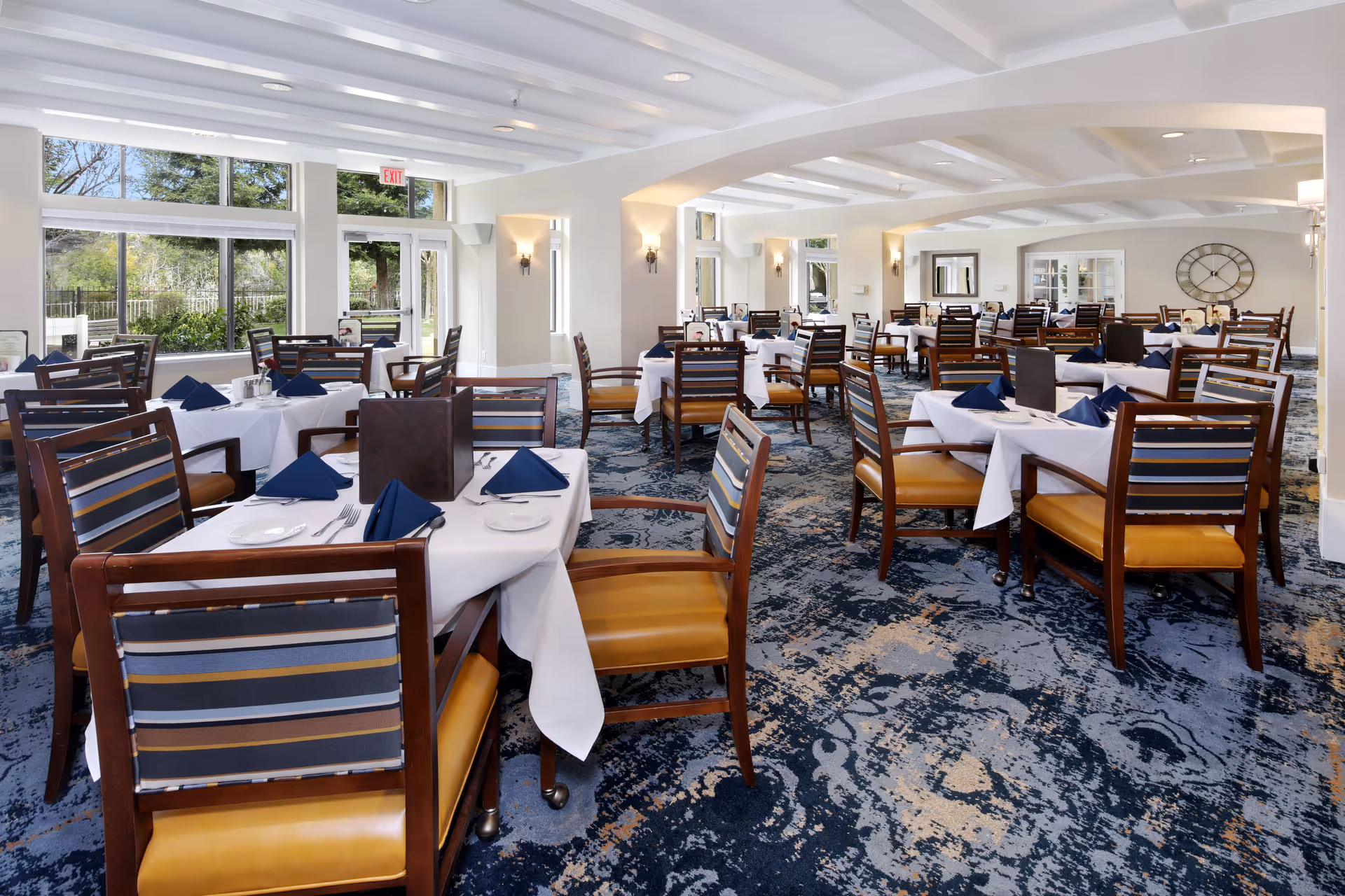 A spacious dining room with multiple tables covered in white tablecloths, each set with plates, silverware, and navy blue folded napkins. The chairs have wooden frames with striped fabric backs and mustard-colored seats. Large windows allow natural light to fill the room, and there is a patterned blue carpet on the floor. Wall sconces and a large clock decorate the light-colored walls.