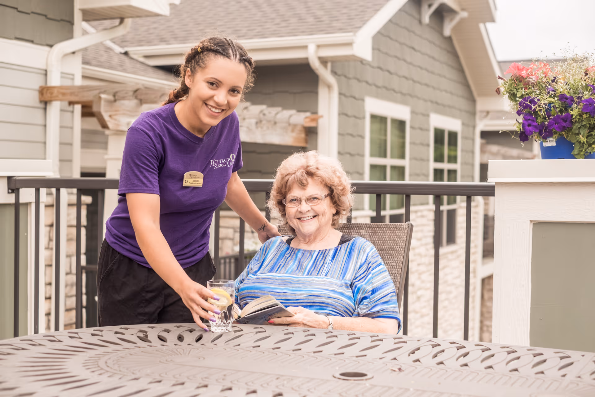 A smiling young female caregiver in a purple Heritage Senior Living shirt serves a glass of water with lemon to a happy elderly woman sitting outdoors on a patio, holding a book. The background shows the exterior of the facility with windows and flower pots.