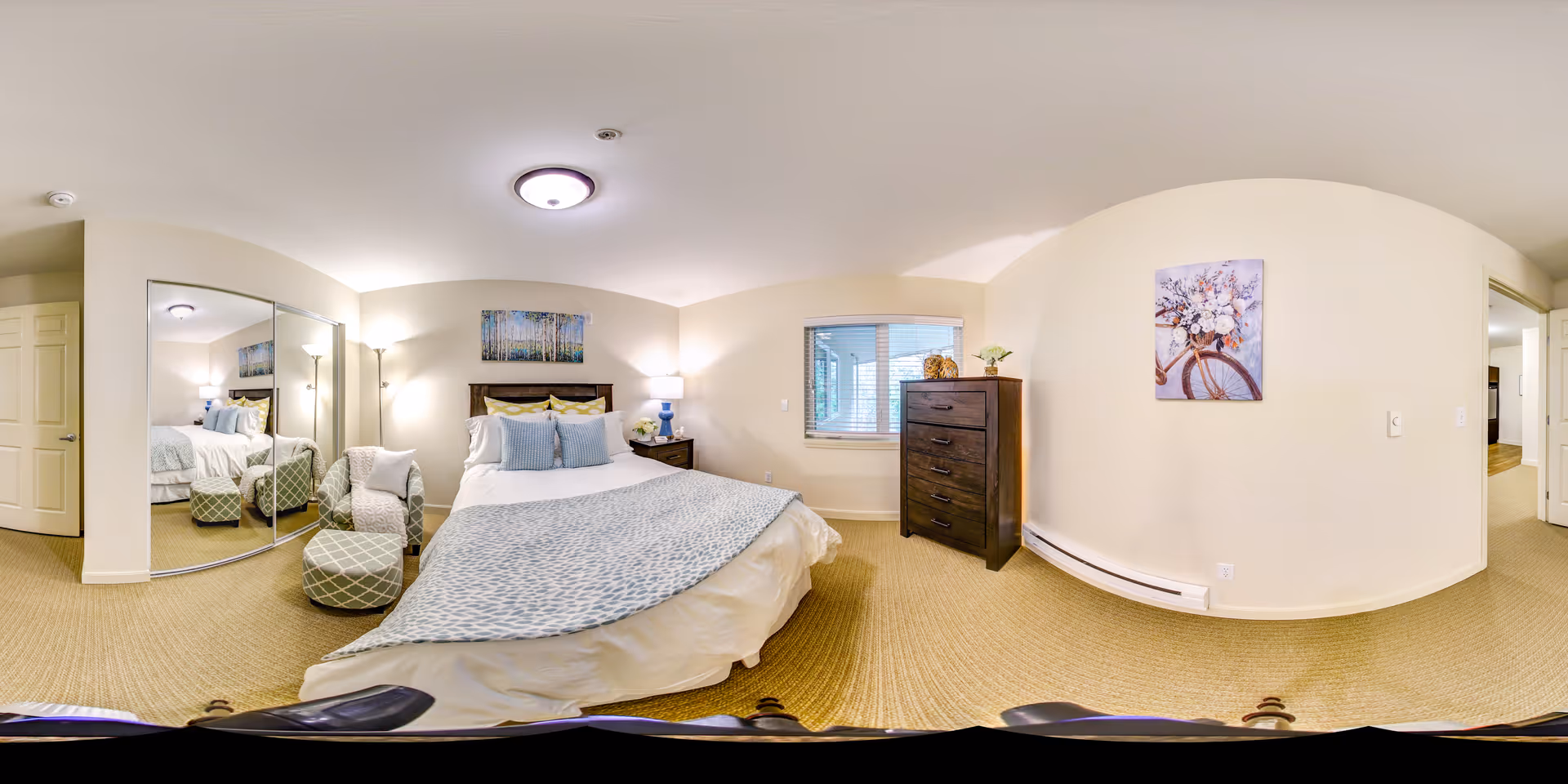 A well-lit bedroom with a large bed covered in white and light blue bedding. There are two bedside tables with lamps, a cushioned chair with an ottoman, a dresser with decorative items, a window with blinds, and a large mirrored closet. The walls are light-colored with two pieces of artwork hanging.