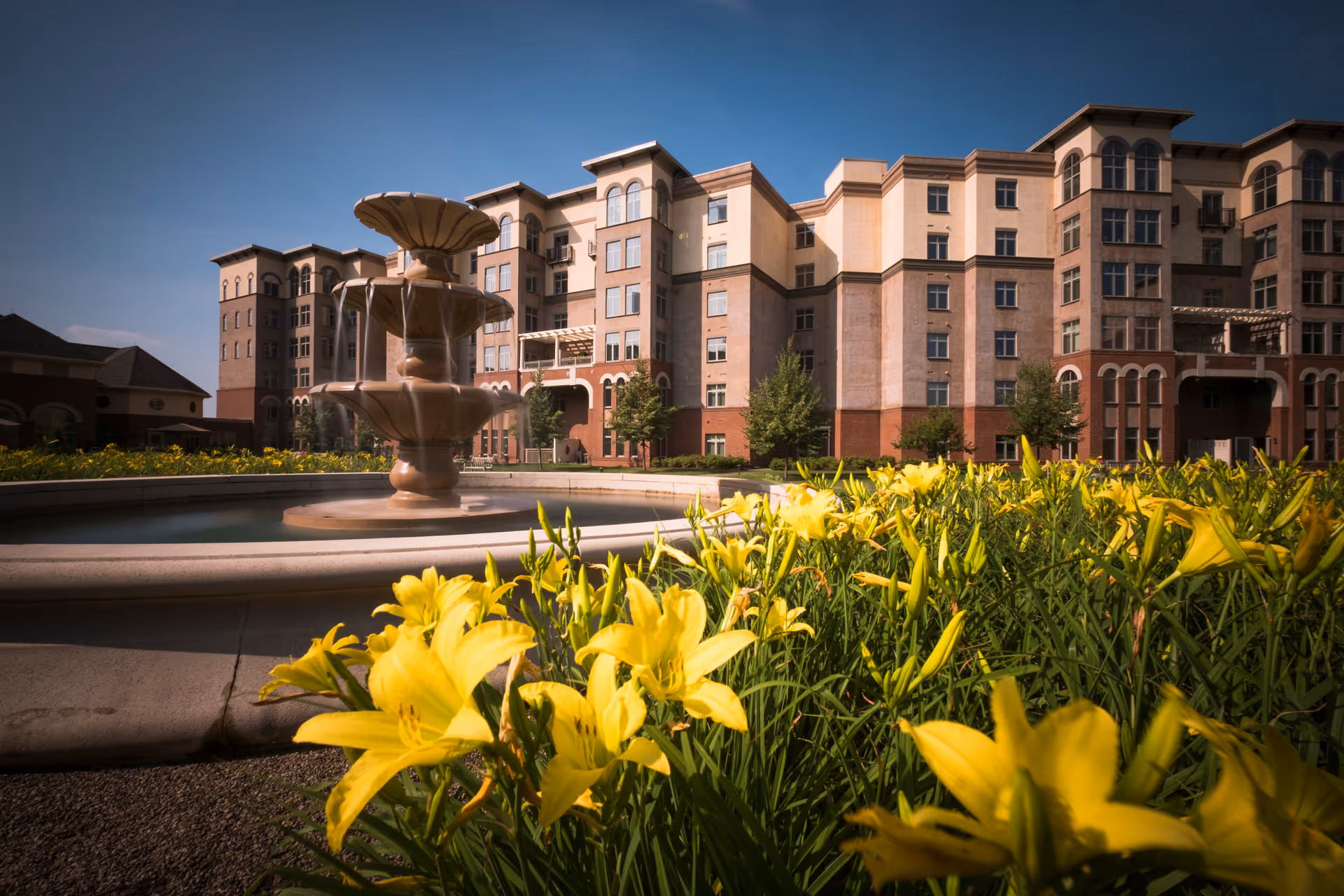 A large multi-story senior living facility building with a beige and brick exterior under a clear blue sky. In the foreground, there is a tiered water fountain surrounded by a circular basin, and bright yellow flowers with green foliage.