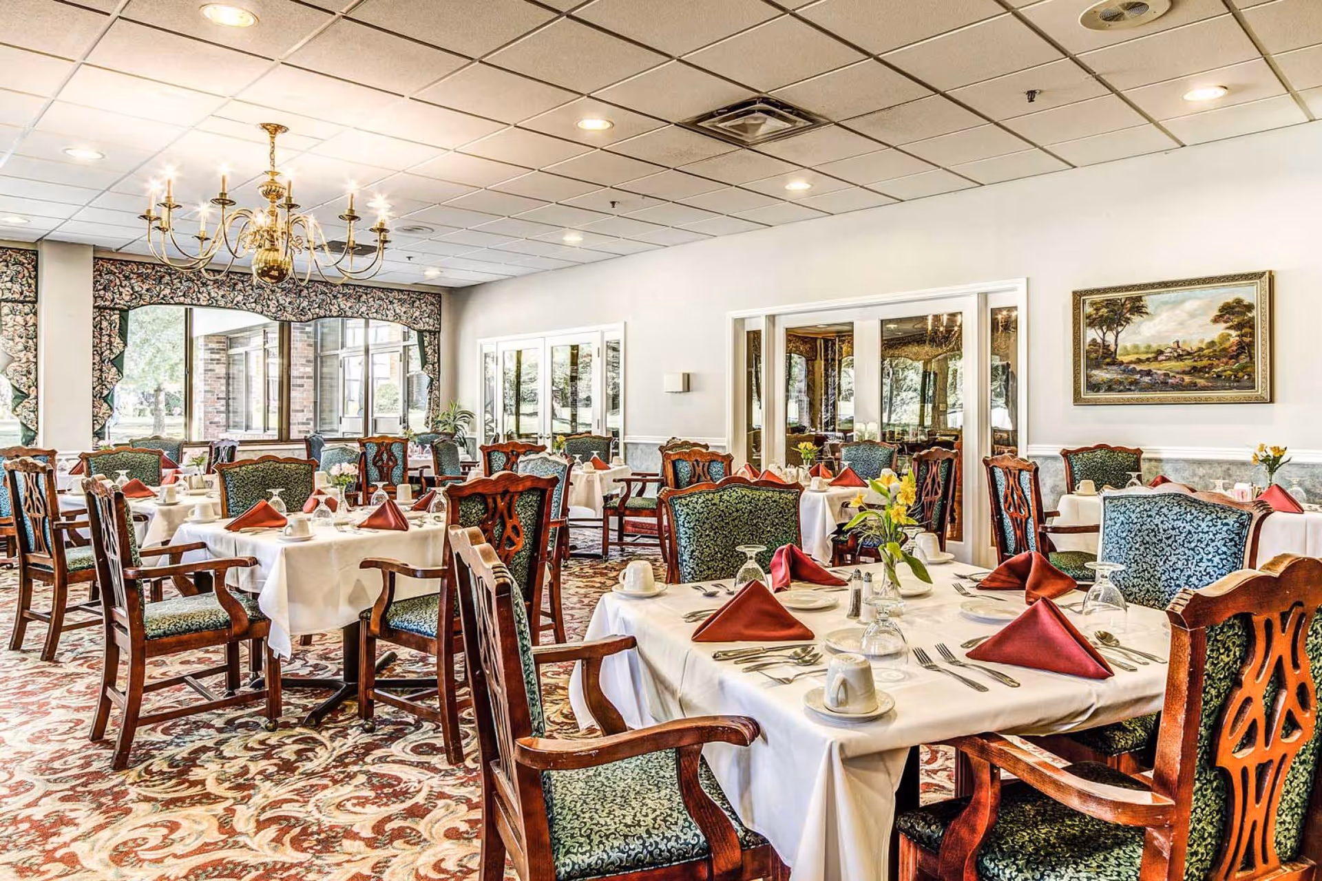 A dining room with multiple tables covered in white tablecloths, each set with red folded napkins, cups, glasses, and silverware. The chairs have wooden frames with green patterned upholstery. A large chandelier hangs from the ceiling, and there is a large window with floral curtains letting in natural light. A framed landscape painting is on the wall.