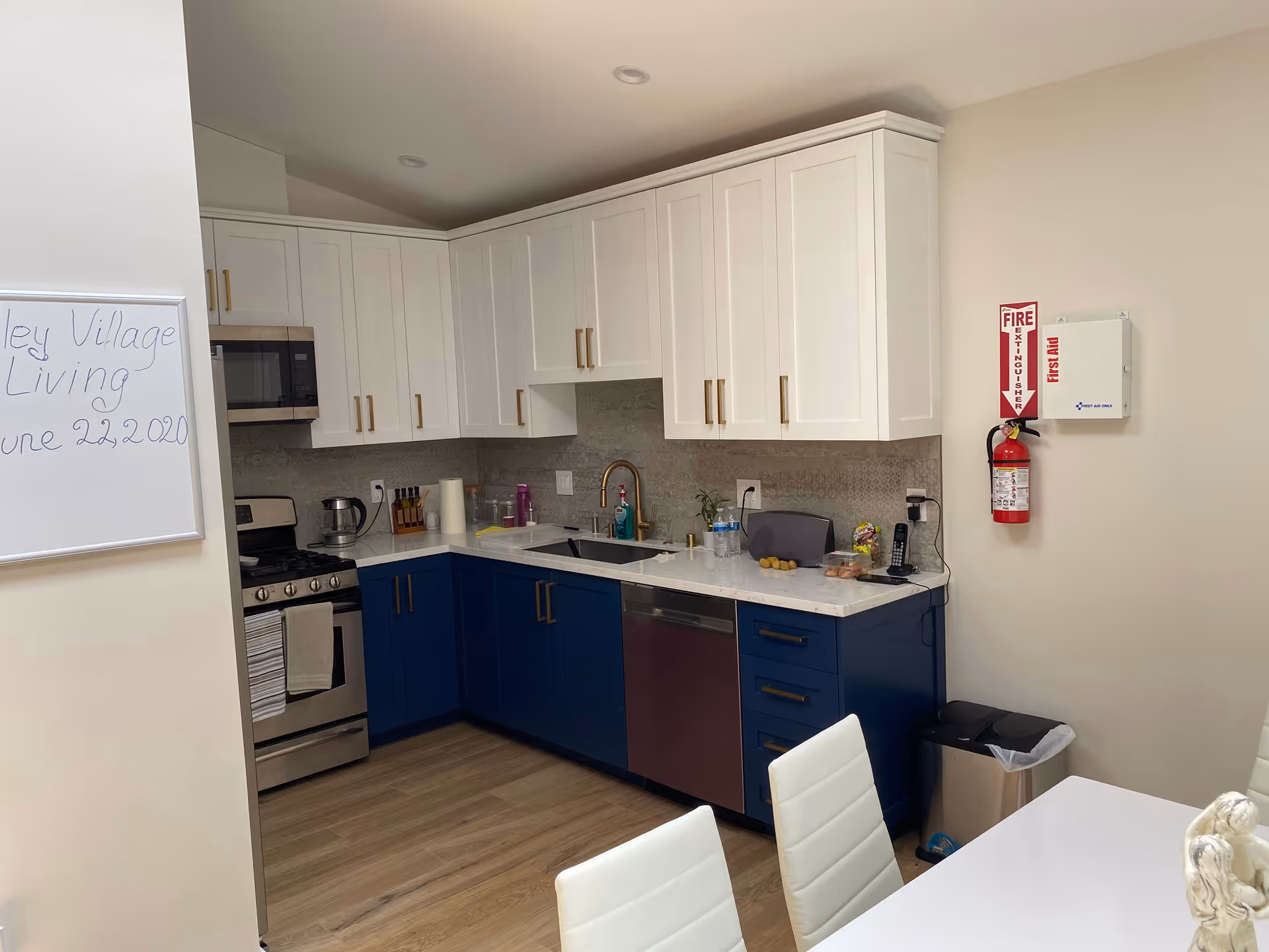 Modern kitchen with white upper cabinets and blue lower cabinets, stainless steel stove and dishwasher, a sink with a gold faucet, and various kitchen items on the countertop. A whiteboard on the left wall reads 'Valley Village Living June 22, 2020'. A fire extinguisher and first aid box are mounted on the right wall. A white dining table with white chairs is partially visible in the foreground.