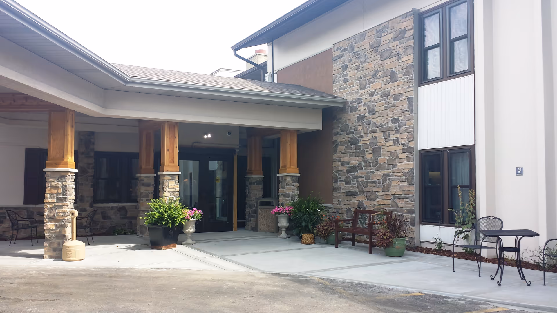 Entrance area of a senior living facility with stone and wood pillars supporting a roof overhang. There are potted plants, a wooden bench, and metal chairs with a small table on the concrete walkway.