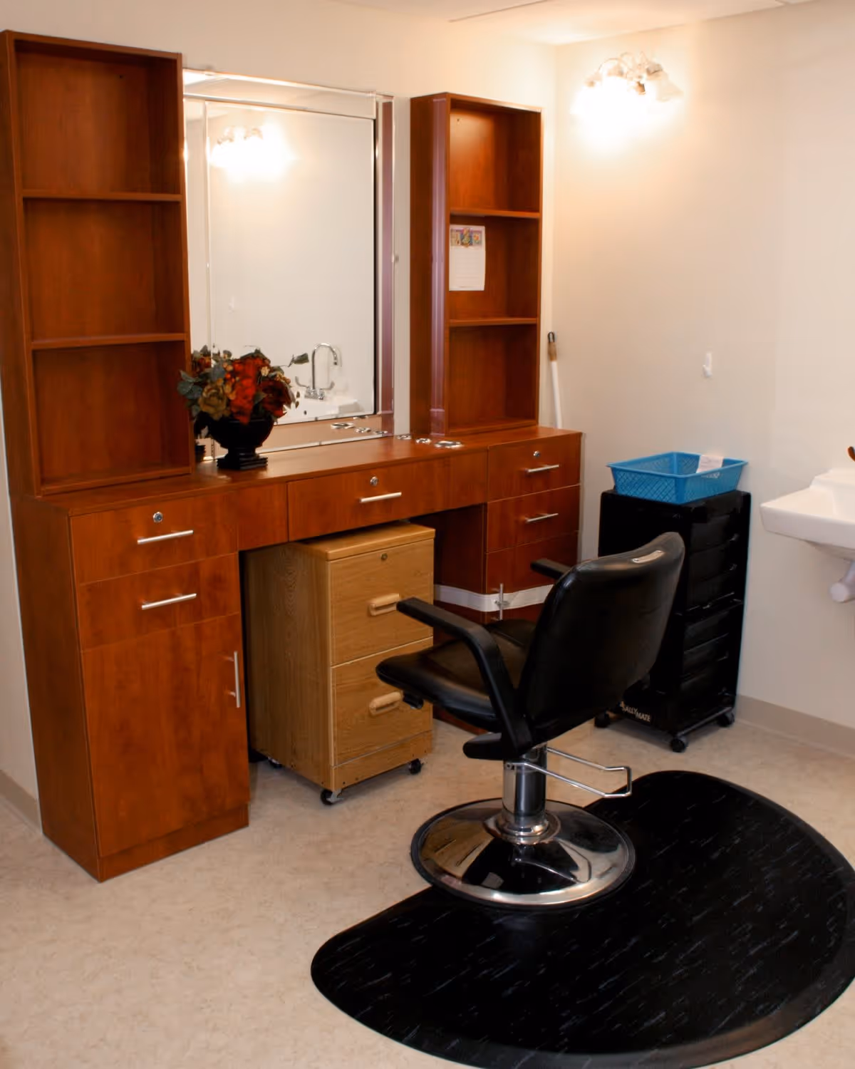 Interior view of a salon area in a senior living facility with a black salon chair on a black mat in front of a wooden vanity with a large mirror. The vanity has shelves, drawers, and a small rolling cabinet underneath. A blue basket sits on a black rolling cart next to a white wall-mounted sink.
