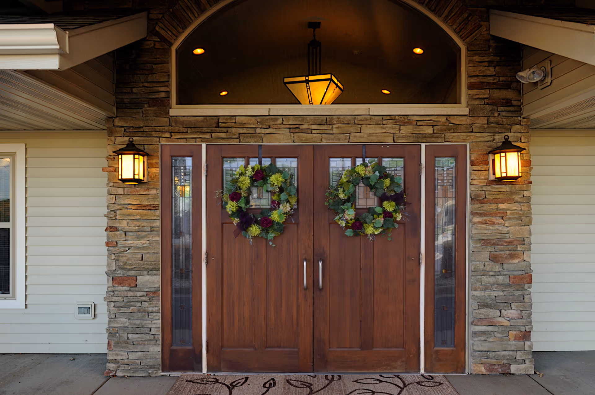 Front entrance of a building with double wooden doors adorned with two green and purple floral wreaths. The entrance is framed by stone walls and two lit lantern-style wall sconces on either side. Above the doors is a large arched window with a hanging light fixture inside. A patterned doormat is placed in front of the doors.