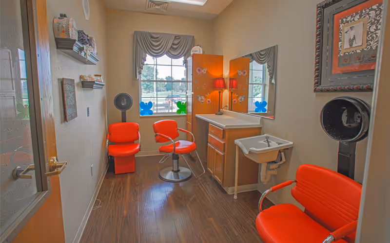 Interior of a small salon or grooming room with three red salon chairs, a sink, a large mirror, a cabinet with a lamp on top, and a window with a valance curtain. The room has wood flooring and beige walls with some decorative items and shelves.
