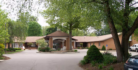 Exterior view of a single-story brick building with a covered entrance, surrounded by trees and greenery, with a driveway and parked cars in front.