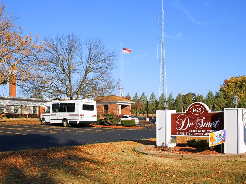 Exterior view of DeSmet Retirement Community showing the entrance sign, a parking lot with a white shuttle bus, a brick building, an American flag on a flagpole, and trees with autumn foliage under a clear blue sky.