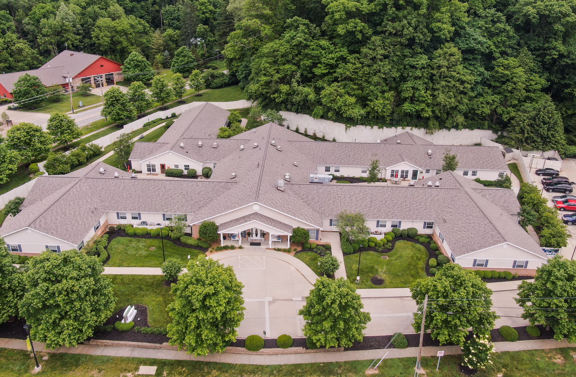 Aerial view of Arden Courts - ProMedica Memory Care Community in Anderson Township, showing a large, single-story building with a gray roof surrounded by green lawns, trees, and landscaped bushes. There is a circular driveway at the front entrance and a parking lot visible to the right. The building is nestled near a wooded area.