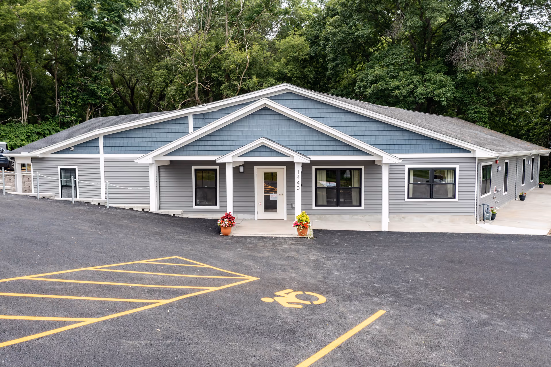 Front exterior view of a single-story assisted living facility building with gray and blue siding, white trim, a small covered entrance, two flower pots with colorful flowers on either side of the entrance, and a handicapped parking space marked in yellow in front. The building is surrounded by trees.