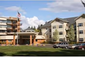 Exterior view of Friendsview Retirement Community showing a multi-story building with beige and brick facade, several windows, an American flag on a flagpole, parked cars, and a green lawn in the foreground under a partly cloudy sky.