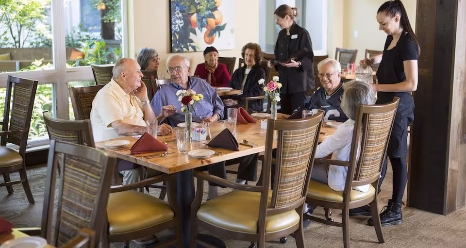 A group of elderly people sitting around a wooden dining table in a well-lit room with large windows. Two staff members are attending to the group, one taking an order and the other pouring a drink. The table is set with plates, glasses, and folded napkins, and there are small flower vases as centerpieces.