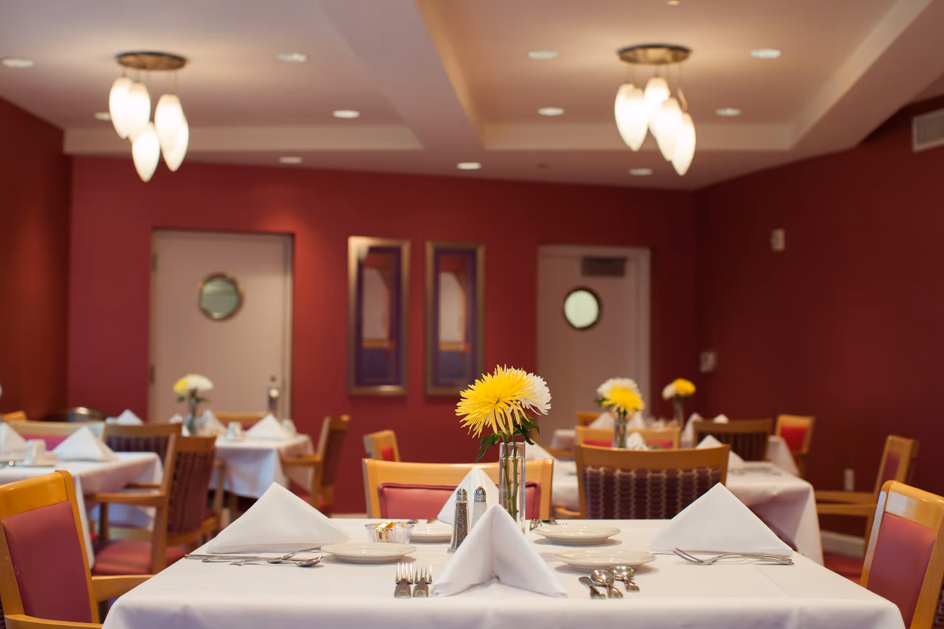 A dining room with tables covered in white tablecloths, each set with plates, silverware, folded napkins, and small vases holding yellow and white flowers. The walls are painted a deep red, and there are two doors with round windows in the background. Ceiling lights with multiple bulbs hang above the tables.