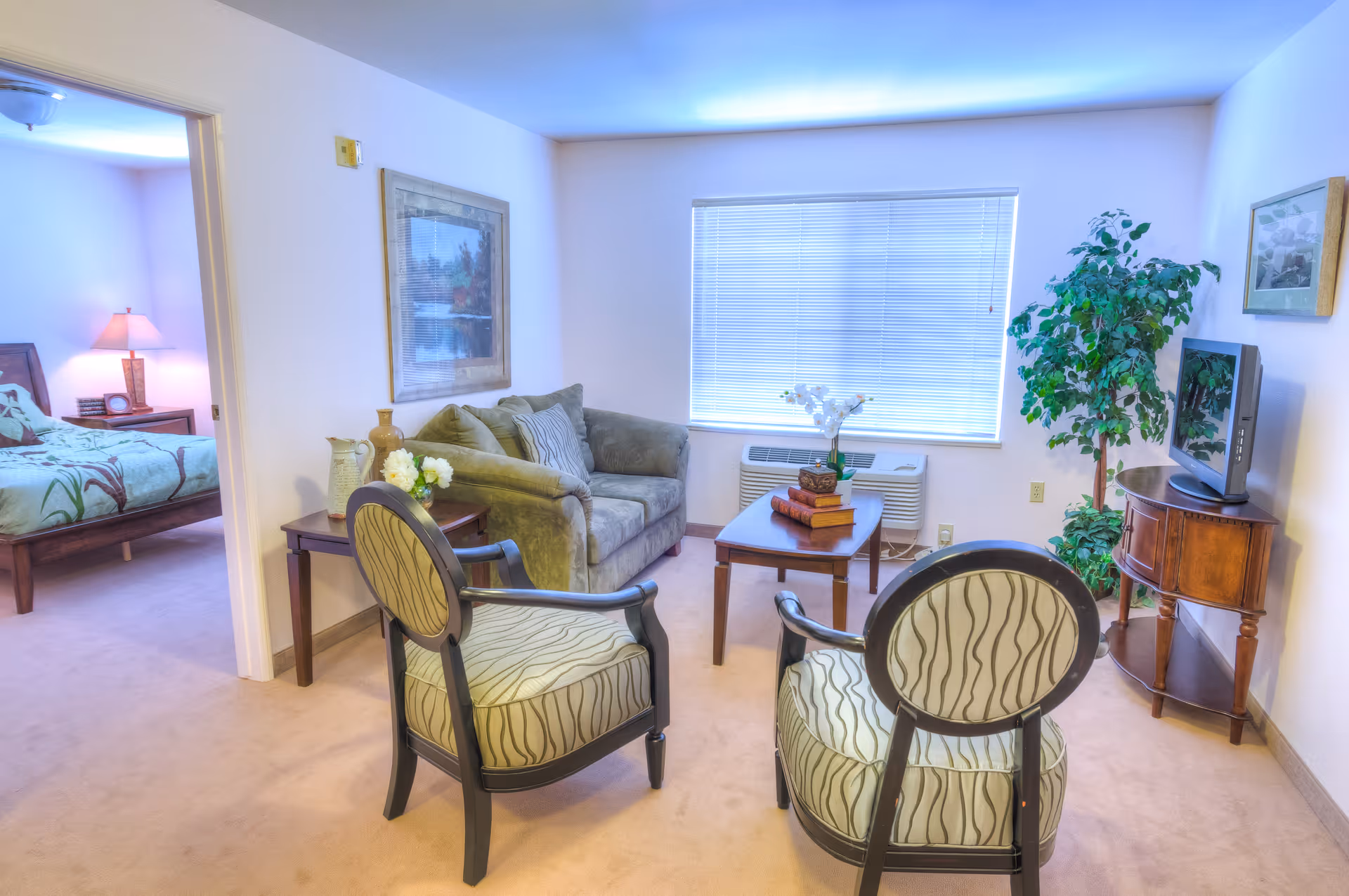 A cozy senior living room with two patterned armchairs facing a green sofa. A wooden coffee table with books and a flower vase sits in front of the sofa. A large window with blinds is behind the sofa. To the right, there is a wooden TV stand with a flat-screen TV and a tall potted plant. A doorway on the left leads to a bedroom with a bed, nightstand, and lamp.