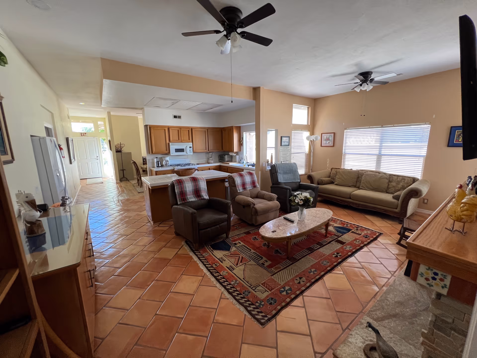 A cozy living room and kitchen area with terracotta tile flooring. The living room features a beige sofa, two armchairs with plaid blankets, a recliner, a patterned area rug, a coffee table with a flower vase, and ceiling fans. The kitchen has wooden cabinets, a microwave, and a white countertop. Large windows with blinds allow natural light to fill the space.