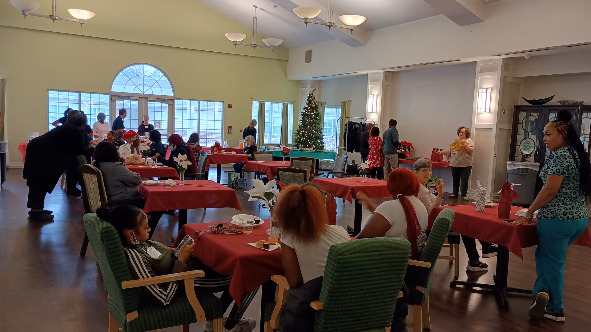 A group of people gathered in a large room with tables covered in red tablecloths. Some people are seated and engaged in conversation, while others are standing. There is a decorated Christmas tree near the windows, and the room has large arched windows letting in natural light. The atmosphere appears to be social and festive.