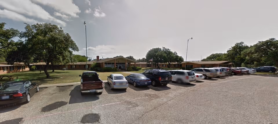 Parking lot with several cars parked in front of a single-story building surrounded by trees under a partly cloudy sky.