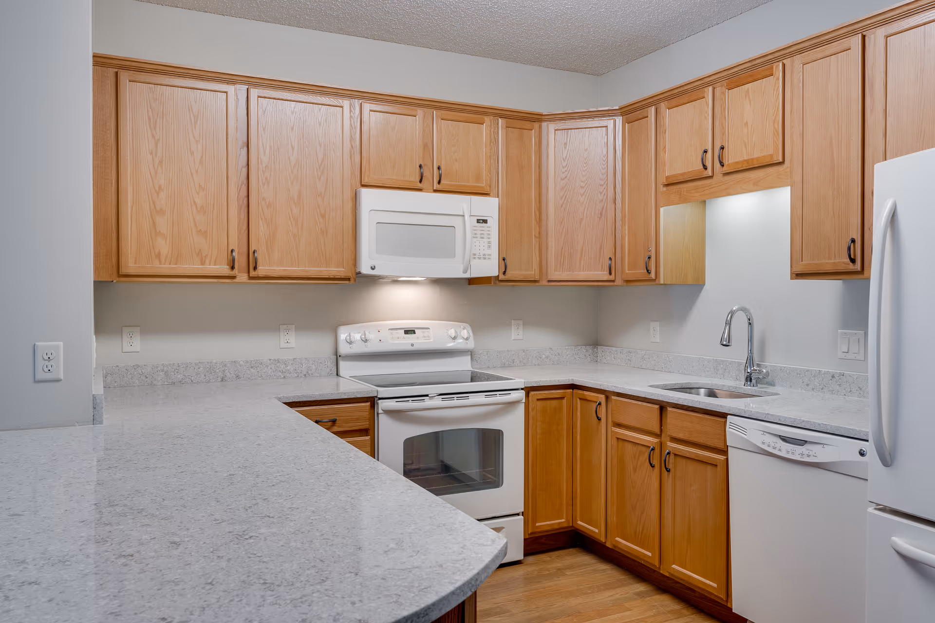 A clean and modern kitchen with light wood cabinets, white appliances including a microwave, oven, dishwasher, and refrigerator, and light-colored granite countertops. The kitchen has a stainless steel sink with a curved faucet and hardwood flooring.