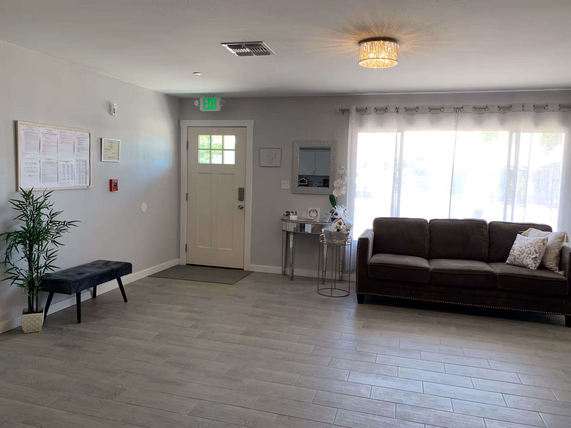 A bright and clean senior living facility common area with a gray tiled floor, a dark gray sofa with decorative pillows near a large window covered with sheer white curtains, a small round side table with a flower arrangement, a rectangular mirror on the wall, a white entrance door with a window, a black bench, a potted plant, and a bulletin board with notices on the wall.