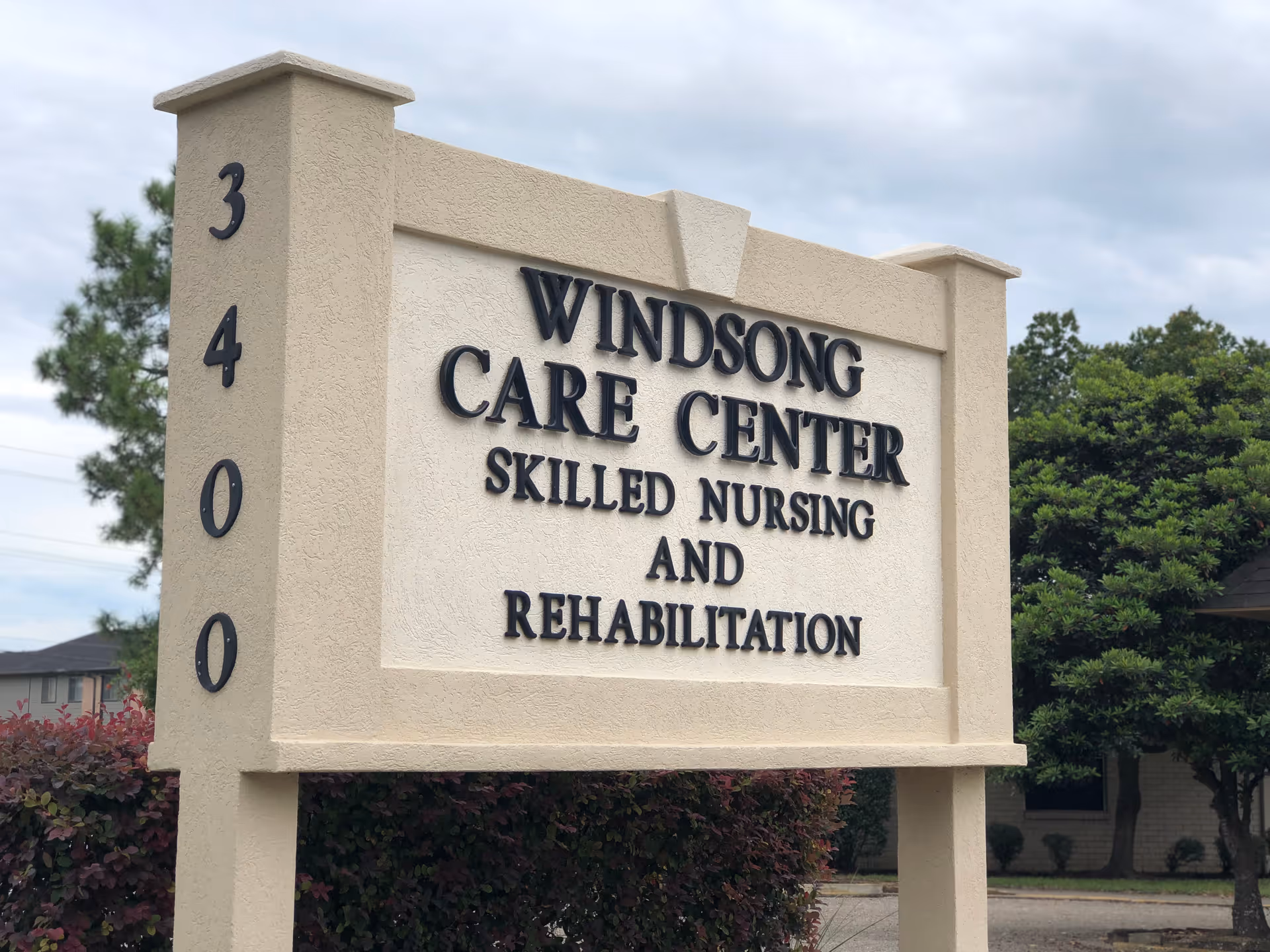 Outdoor sign for Windsong Care Center, a skilled nursing and rehabilitation facility, with the address number 3400 on the side. The sign is beige with black lettering and surrounded by greenery and trees under a cloudy sky.
