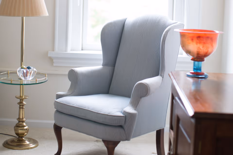 A light blue upholstered wingback chair positioned near a window with white trim. To the left of the chair is a brass floor lamp with a glass side table holding a decorative glass object. To the right is a wooden piece of furniture with a red and blue decorative glass bowl on top.