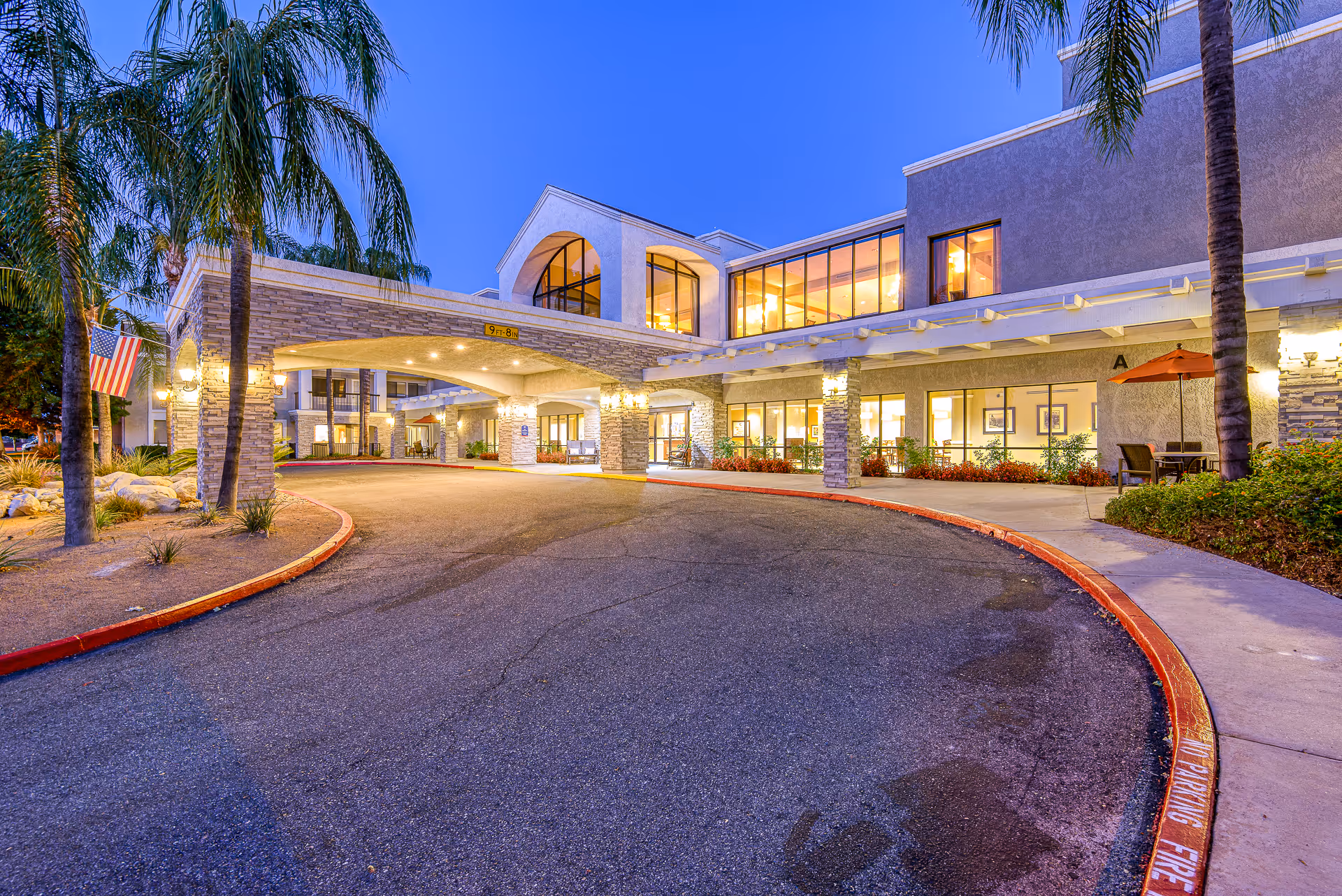 Exterior view of Menifee Senior Living facility at dusk, showing a covered entrance with stone pillars, large windows with warm interior lighting, palm trees, and an American flag on the left side.